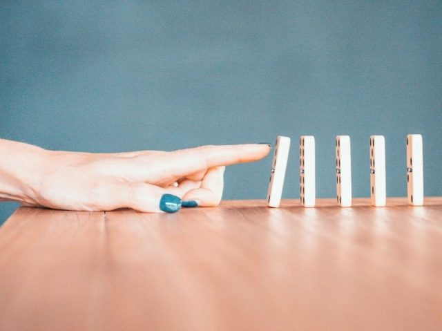 Mão tocando peças de dominó, simbolizando o início de um processo e o impacto de cada etapa no atendimento ao cliente
