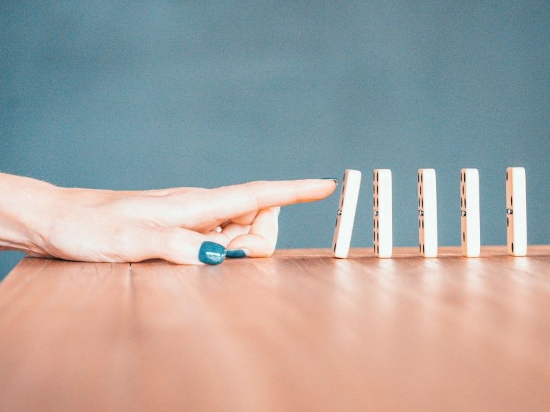 Mão tocando peças de dominó, simbolizando o início de um processo e o impacto de cada etapa no atendimento ao cliente