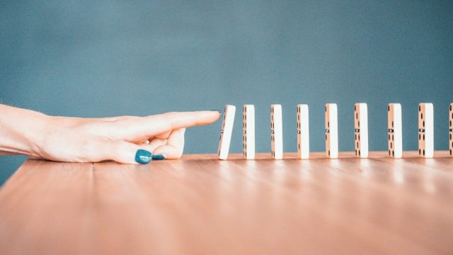 Mão de uma pessoa com unhas pintadas de azul interrompendo a queda de uma fileira de peças de dominó sobre uma mesa de madeira. A imagem transmite a ideia de prevenção, controle de danos ou interrupção de uma sequência de eventos. Ao fundo, há um fundo azul sólido desfocado.