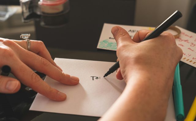 Mãos escrevendo em um envelope branco com a palavra “To:” usando uma caneta preta, simbolizando o envio de uma mensagem personalizada — metáfora para a criação de uma carta de vendas direcionada ao cliente certo. Perguntar ao ChatGPT
